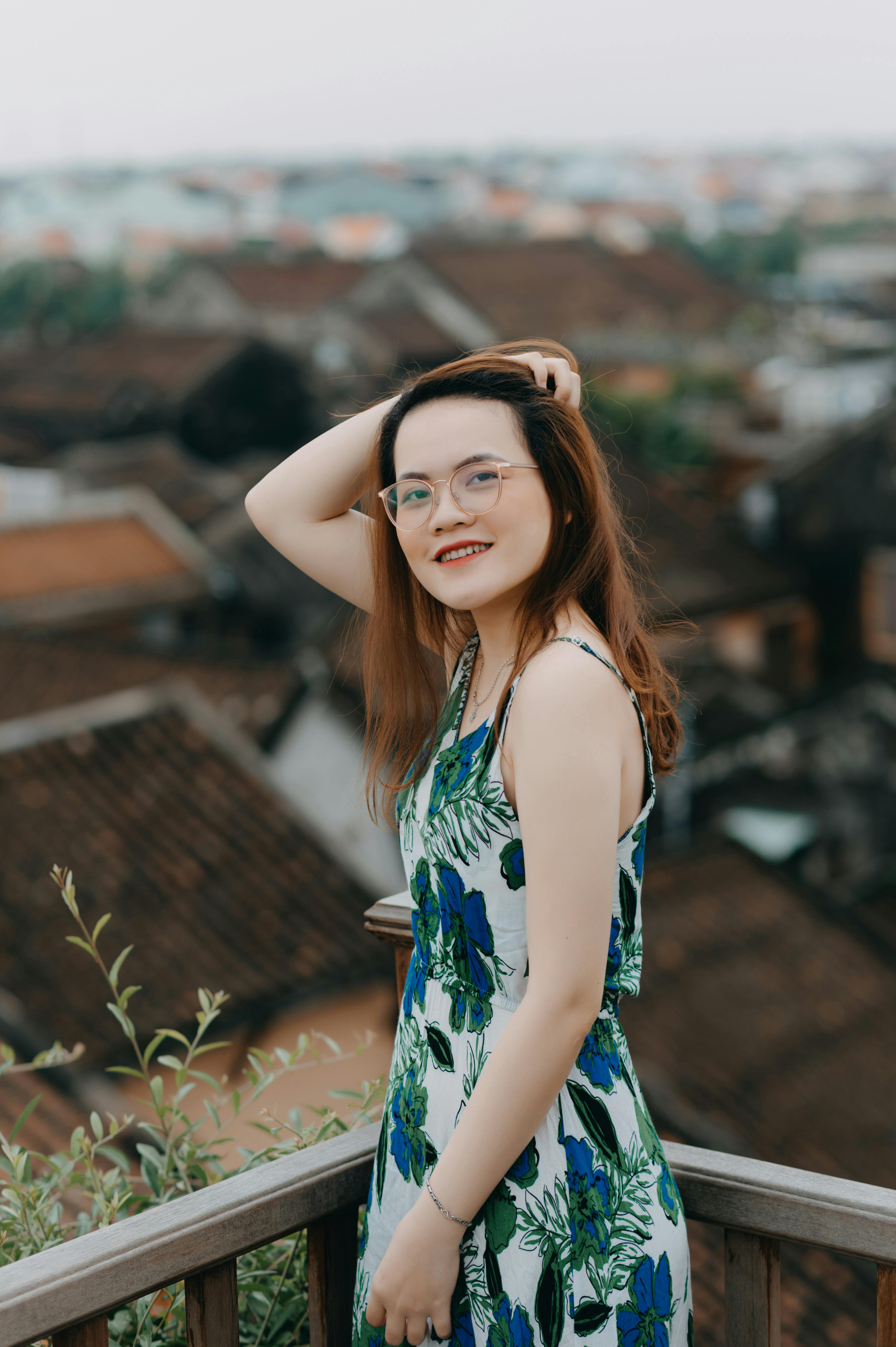 Happy Asian female in gentle dress and spectacles touching hair and looking at camera while spending time on balcony in overcast day