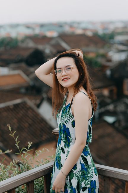 Happy Asian female in gentle dress and spectacles touching hair and looking at camera while spending time on balcony in overcast day