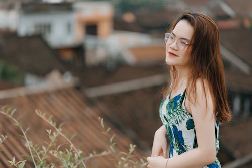 Side view of dreamy young Asian female traveler in stylish dress and eyeglasses standing on viewpoint and admiring old town during sightseeing trip