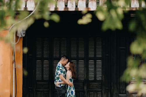 Free Side view of loving Asian couple hugging while standing near building in countryside with green leaves during date on summer day Stock Photo