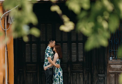 Side view of tender Asian boyfriend kissing forehead of girlfriend while cuddling near building in countryside with green leaves during date