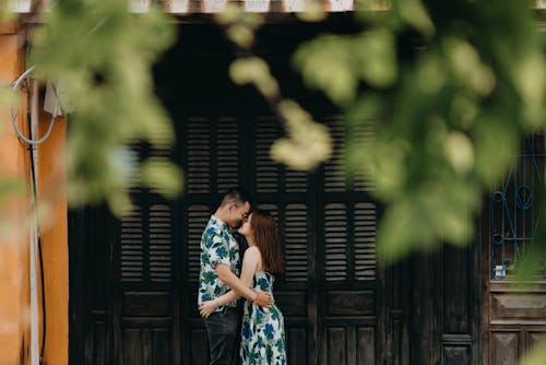 Side view of romantic Asian couple cuddling while standing near building in rural area with green plants during romantic date
