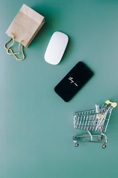 Top view of a smartphone, mouse, paper bag, and mini shopping cart on a green background, symbolizing e-commerce.