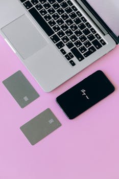 Image of a laptop, credit cards, and smartphone on a pink background, symbolizing online shopping.