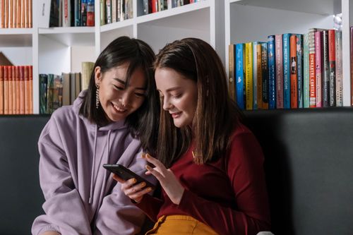 Free Two teenage girls sitting in a library sharing a mobile phone, smiling and enjoying their time. Stock Photo