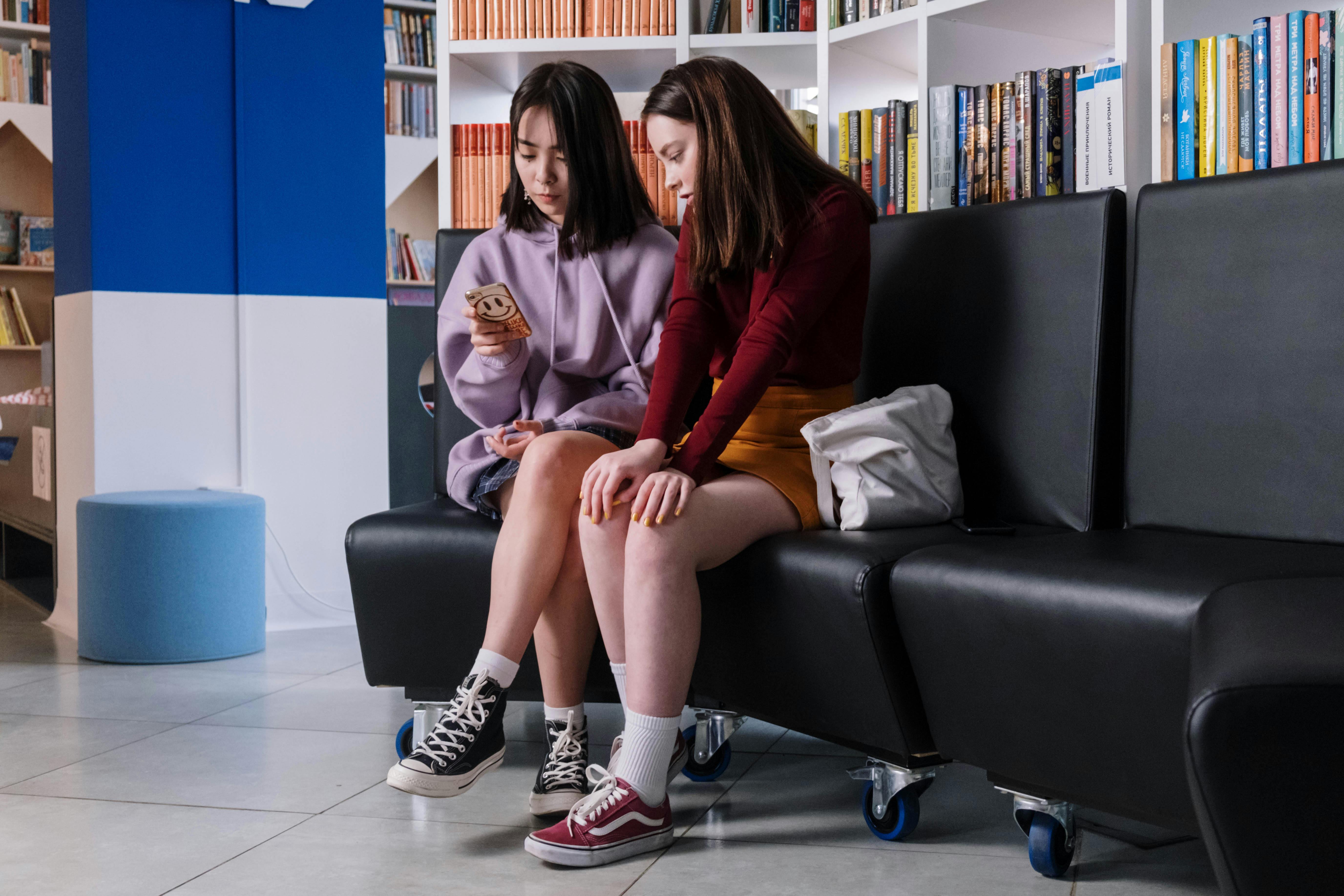 two teenagers sitting on a couch looking at a smartphone