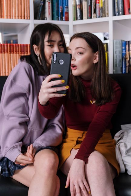 Two teenage girls sitting indoors, capturing a selfie together with a smartphone.