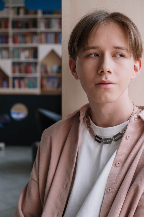 Free Teenager standing near bookshelf in a modern library, looking thoughtful. Stock Photo