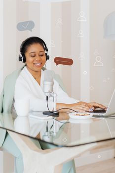 Young woman recording a podcast at home using a laptop and microphone, smiling and engaging with audience.