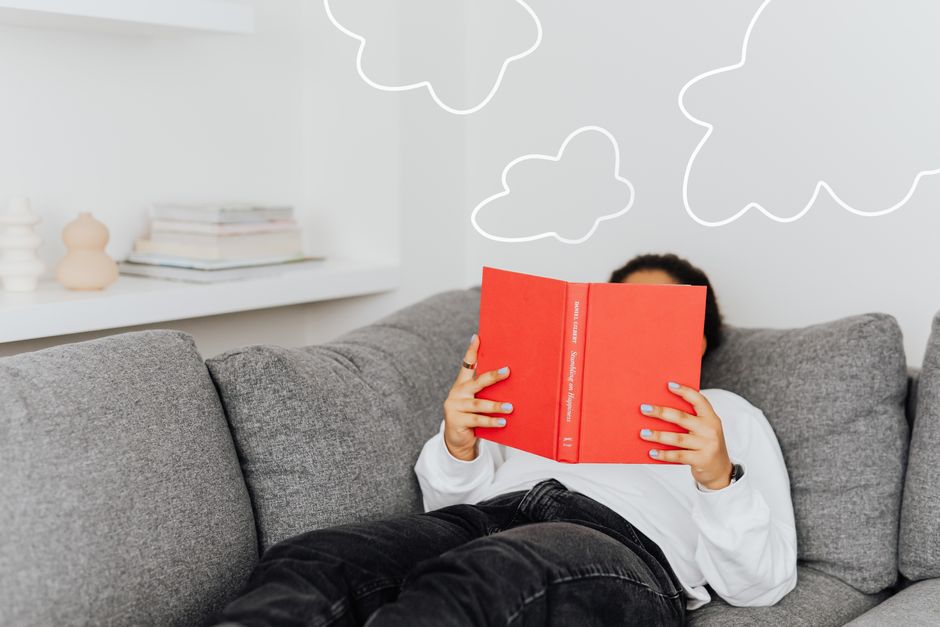 Person relaxing with a book on a sofa. Drawn clouds add a whimsical touch.