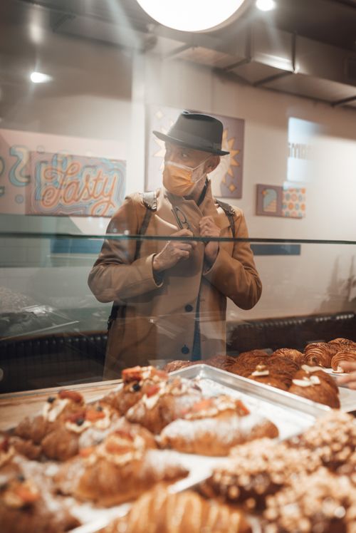 Free Man wearing beige coat and hat and medical mask standing beside glass showcase in bakery shop and looking away Stock Photo