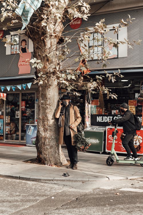 Full body of male in outerwear and mask with bouquet of branches talking on mobile phone near tree and man riding electric scooter