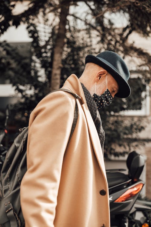 Side view of old male in stylish hat and coat with backpack protecting face from coronavirus with mask