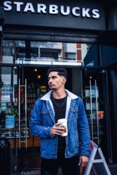 Young man with a coffee cup stands confidently outside a cafe in a stylish denim jacket.