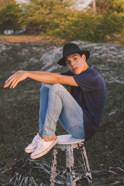 Side view full length of young man in jeans and sneakers with hat and t shirt looking at camera while sitting on chair on ground near green bushes in summer day