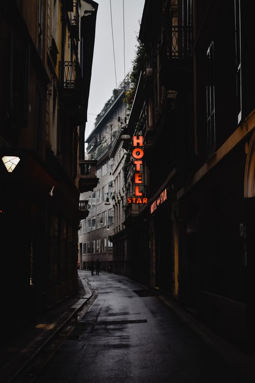 Free Dark, narrow street in Milan featuring a glowing neon hotel sign, creating a moody urban atmosphere. Stock Photo