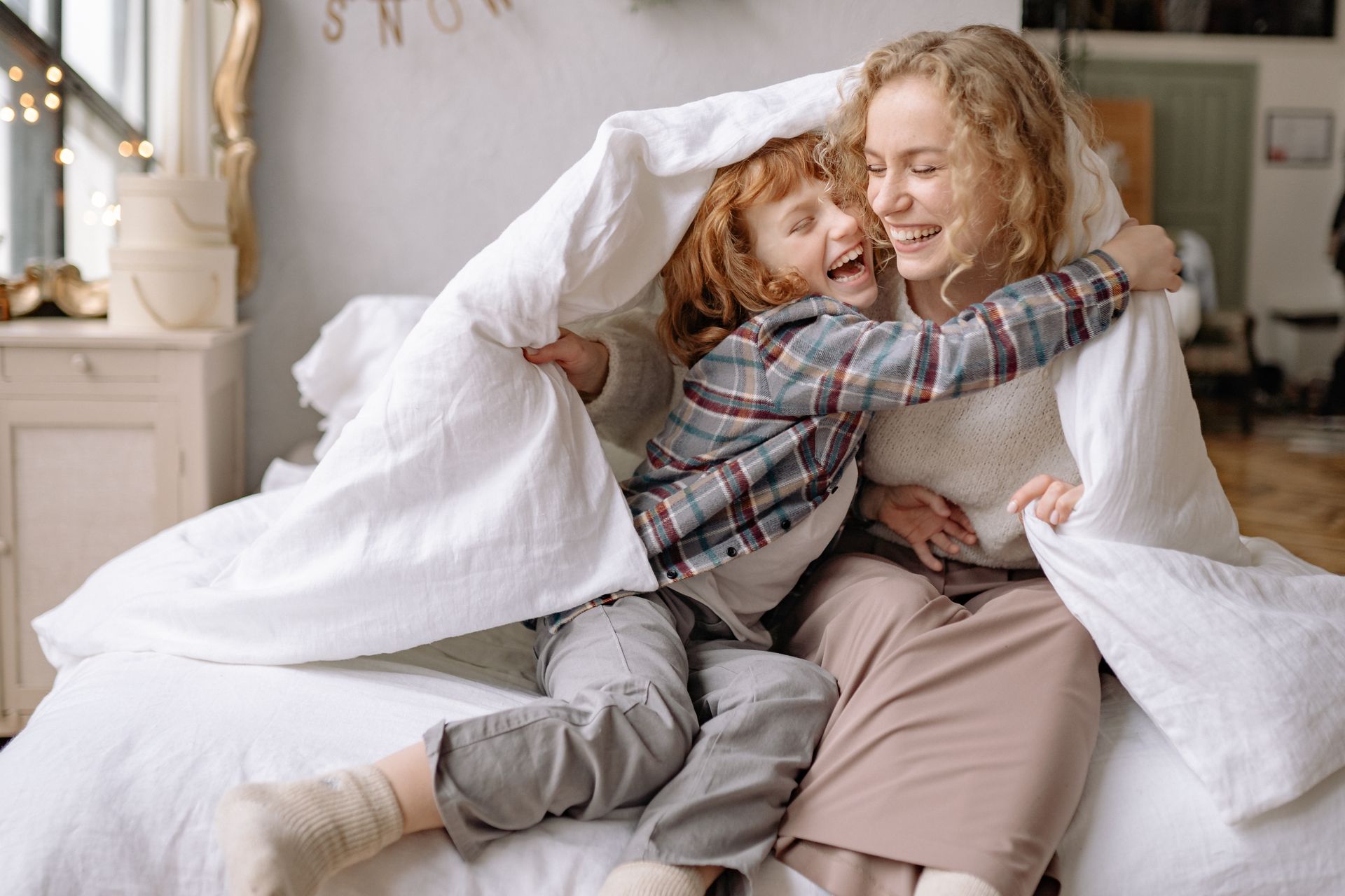 A mother and daughter share a joyful moment, wrapped in a cozy blanket at home.