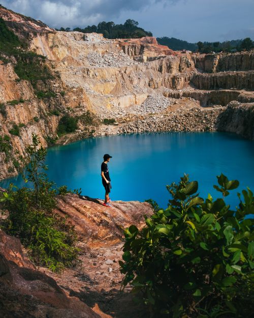 Free A lone adventurer stands by a striking blue lake in a rocky quarry, surrounded by nature. Stock Photo