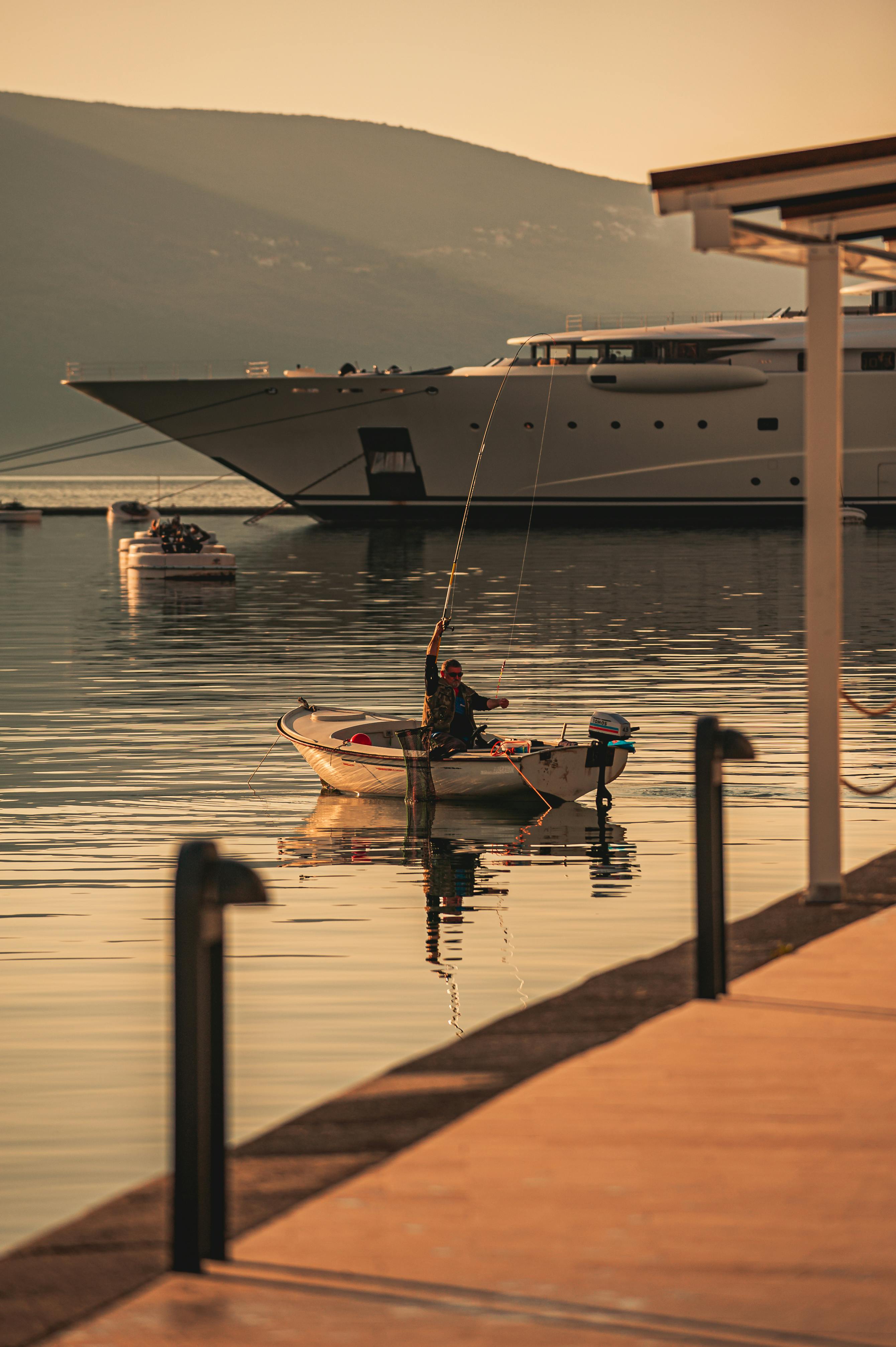 Free A fisherman in Tivat, Montenegro, casts his line at sunrise with luxury yachts in the backdrop. Stock Photo