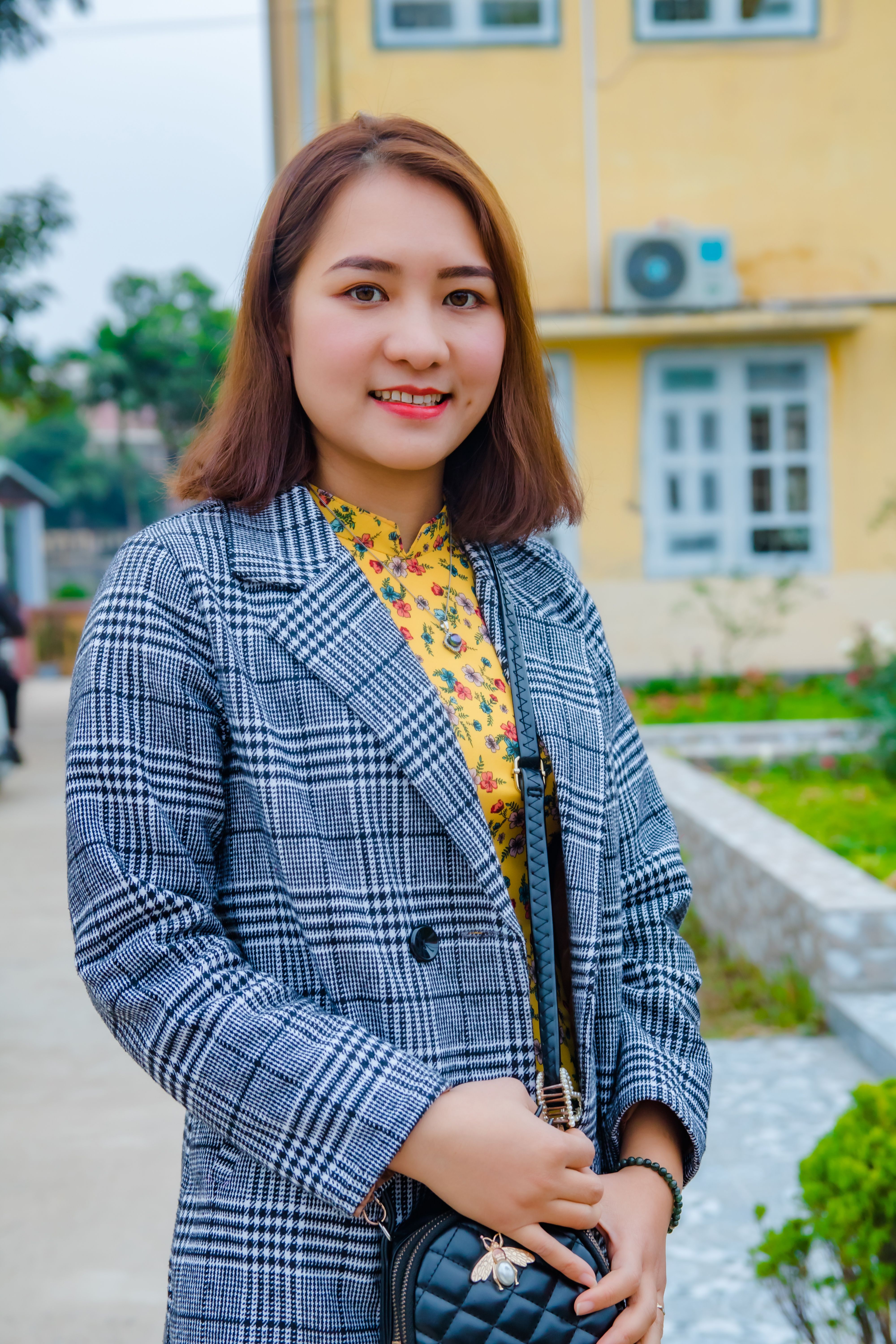 Young woman in plaid blazer smiling outdoors in a vibrant setting.