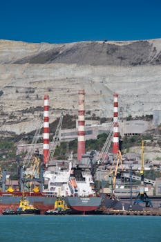 Cargo ship at an industrial port with cranes and smokestacks against a rocky backdrop.