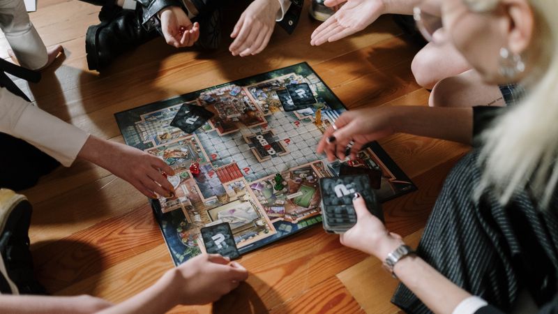 Mehrere Hände um ein aufgeklapptes Brettspiel mit Karten und Spielfiguren auf einem Holzboden