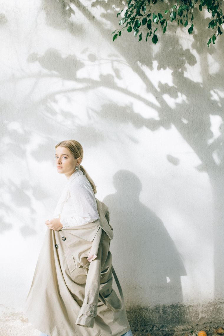 Woman in a beige coat walking past a sunlit wall — photo by Nataliya Vaitkevich on Pexels