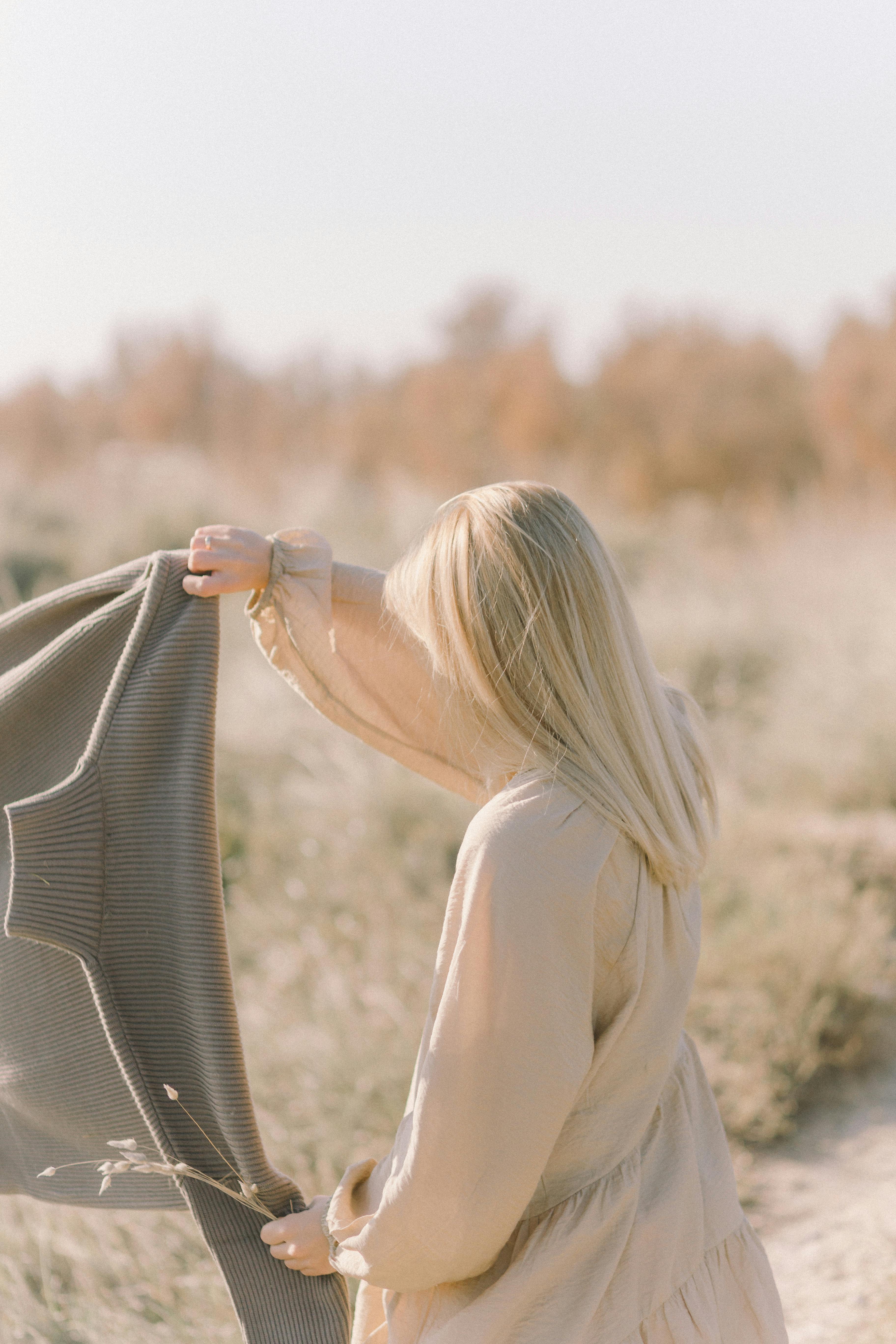 Blonde woman holds a sweater in a serene outdoor setting with autumn colors.