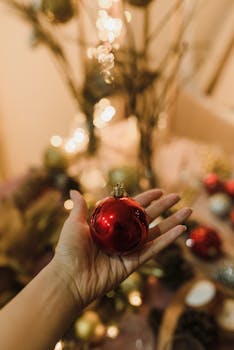 A red Christmas ornament held in a hand with a blurred festive background.