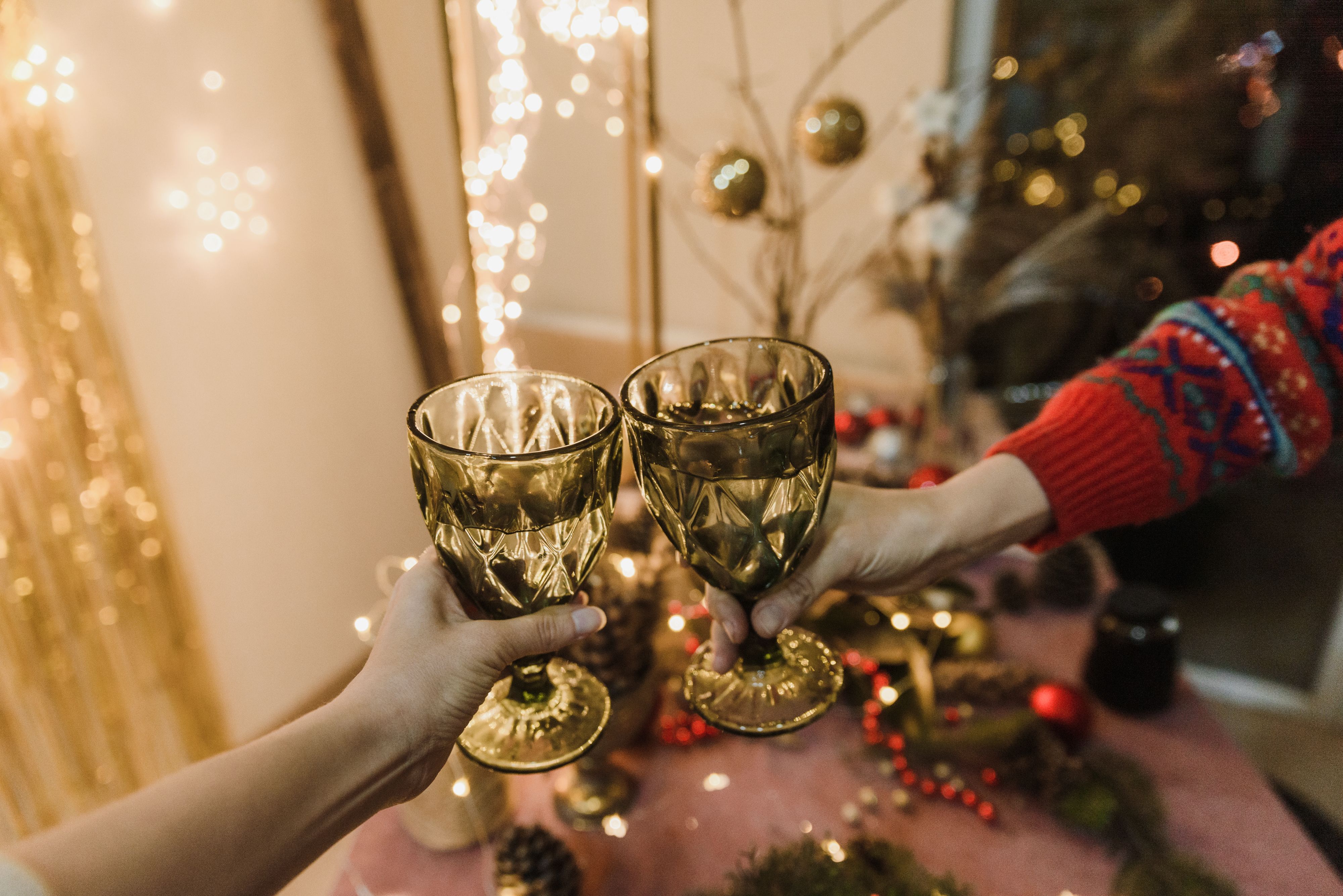 Two hands clinking glasses in a warm festive setting with holiday decorations and lights.