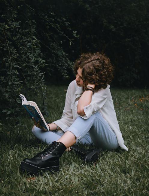 Full length of serious young female in casual clothes sitting on green grassy meadow near plants and bushes while reading book in summer day