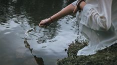 Anonymous woman with flower touching pond on grassy shore