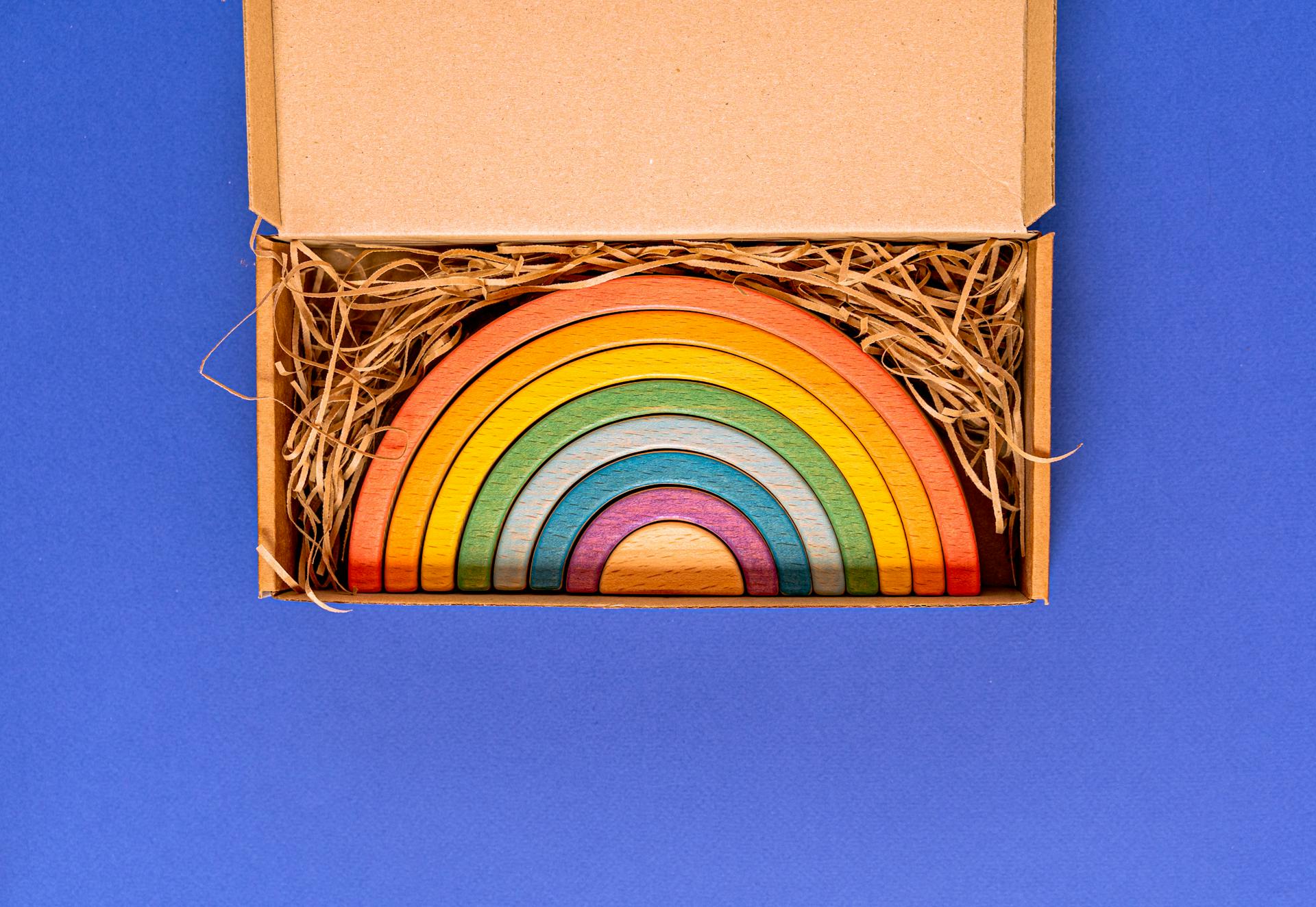 Vibrant wooden rainbow toy packaged in a cardboard box with hay on a blue background.