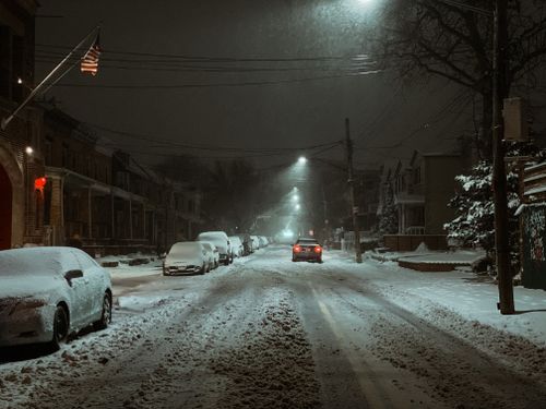 Free Snowy road with parked vehicles near buildings and streetlights in twilight under dark sky in city district in winter Stock Photo