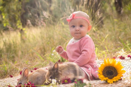 Cute baby sitting with rabbits and sunflowers in a sunny outdoor setting.