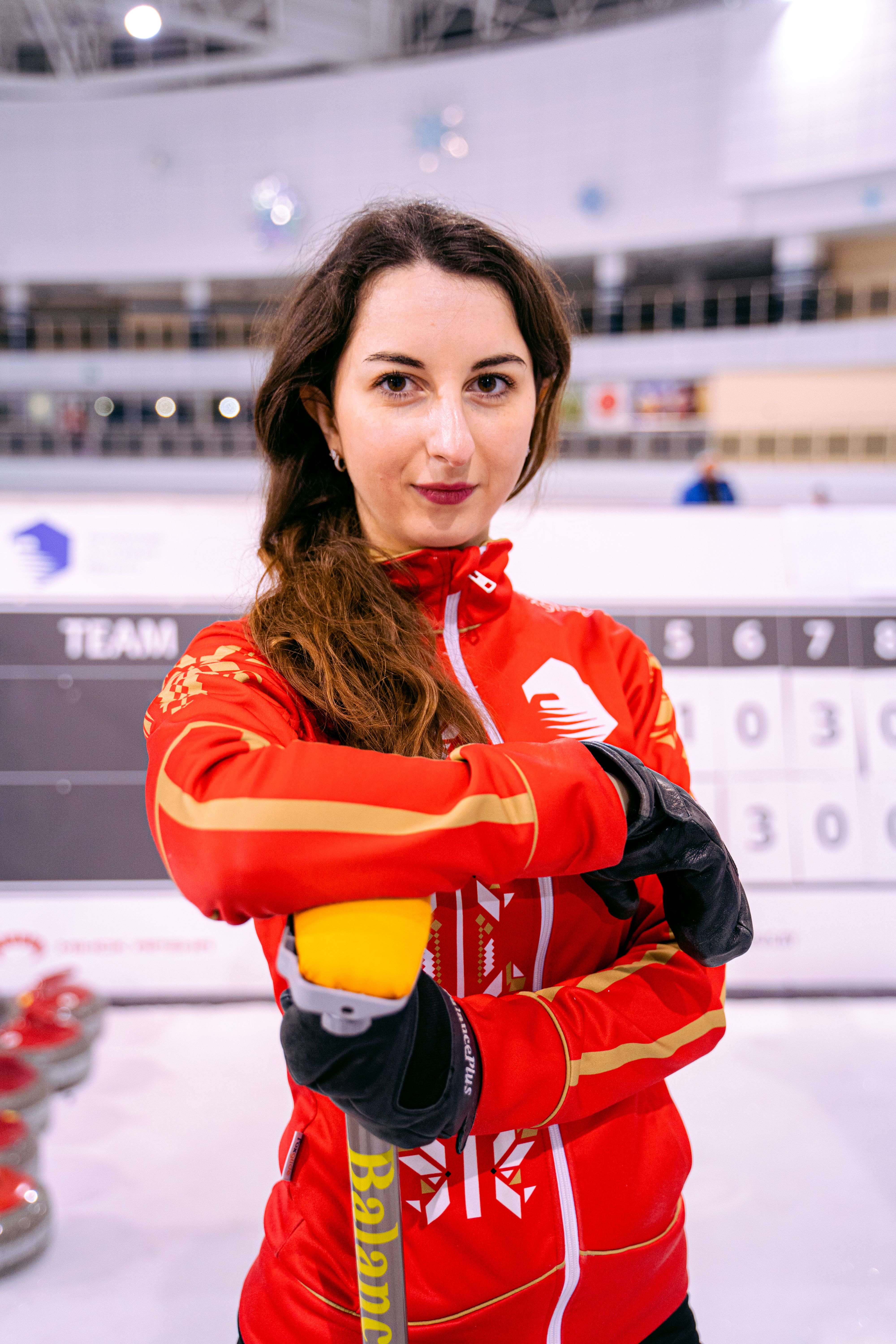 Confident female athlete in red jacket ready to play curling indoors.