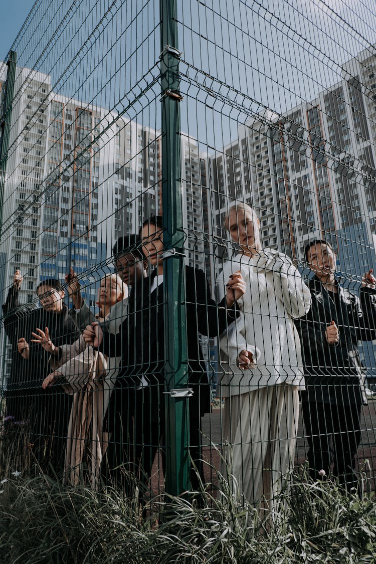 A Group Of Young People Standing Behind A Fence In City 