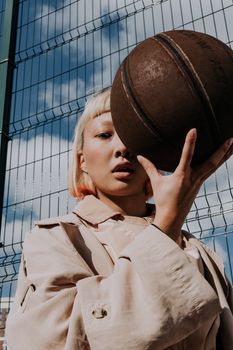 Blonde woman poses with basketball against a fence outdoors, reflecting urban style.
