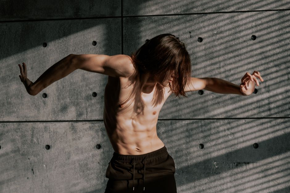 Shirtless male dancer striking a pose in dramatic lighting against a textured wall.