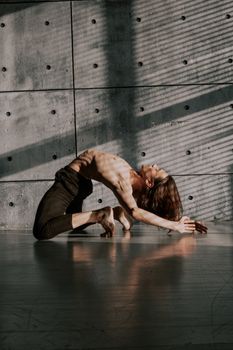 A male dancer with long hair performs a dramatic pose against a textured concrete wall.