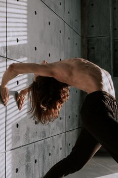 A shirtless dancer stretches dramatically against a textured concrete wall in moody lighting.