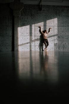 Shirtless dancer leaning against a concrete wall with raised hands in dramatic lighting.
