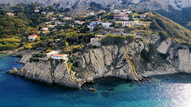 Drone shot of houses on a coastal cliff surrounded by lush greenery and clear blue sea.