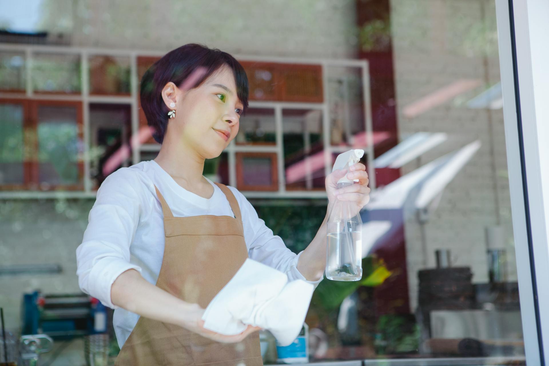 Asian woman cleaning a glass window in a small business environment with a spray bottle and cloth.