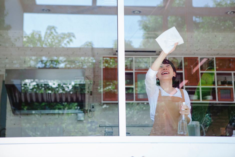 Photo by Antonius Ferret on Pexels Asian woman in apron cleans window with spray bottle and tissue paper, smiling joyfully.