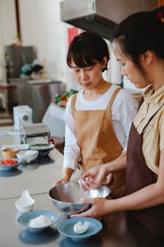 Two Asian women baking together in a modern kitchen, preparing dough with fresh ingredients.