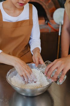 Two people preparing dough by mixing flour and water in a stainless steel bowl indoors.
