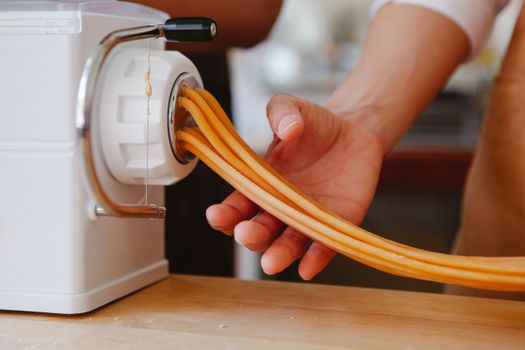 Hand operating a pasta maker extruding fresh, uncooked noodles, close-up view.