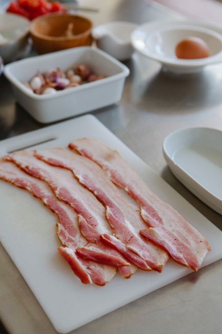 Vacuum-sealed package of raw bacon strips resting on a refrigerator shelf next to a container of cooked bacon slices.