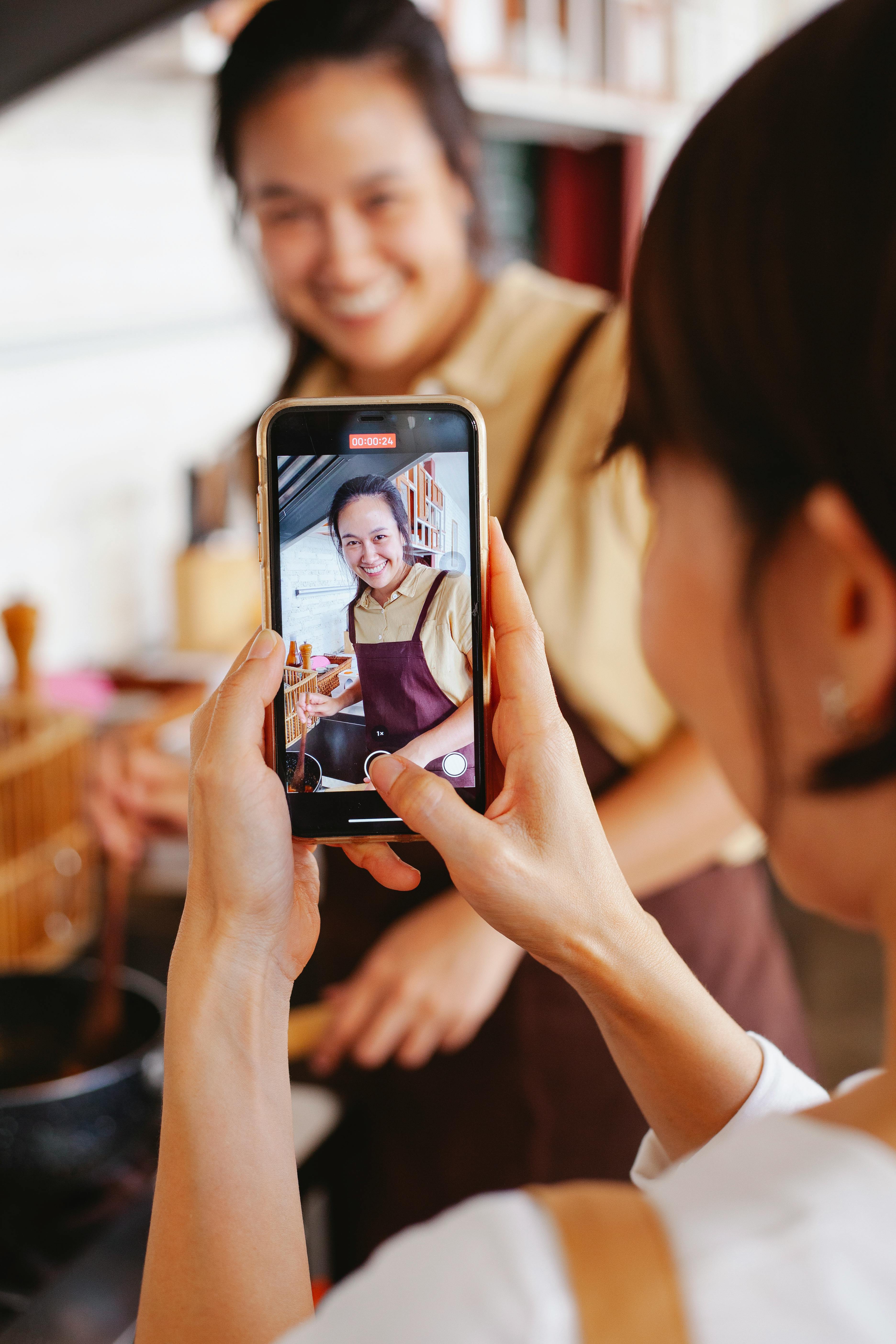 Free Women enjoy cooking together while capturing moments using a smartphone in a modern kitchen. Stock Photo